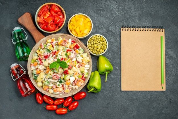 Vista dall'alto della ciotola di insalata di verdure sul supporto del piatto con verdure e bottiglie di olio e aceto e blocco note sul lato e posto per il testo su sfondo grigio scuro