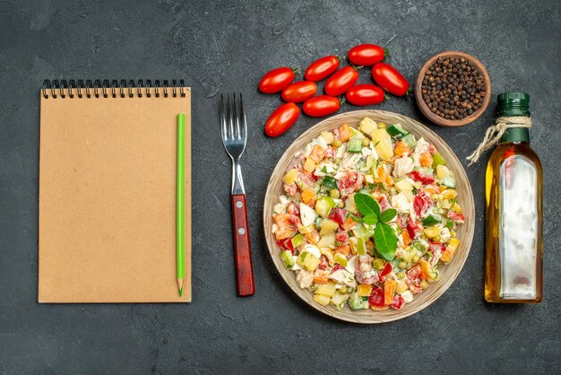 Vista dall'alto della ciotola di insalata vegetariana con forcella olio bottiglia pomodori pepe e blocco note sul lato su sfondo grigio scuro