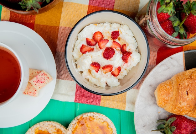 Vista dall'alto della ciotola di ricotta con fragole tazza di tè fette biscottate a mezzaluna rotolo sul panno sulla superficie verde