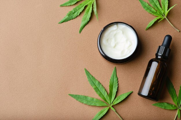 Vista dall'alto della crema cosmetica all'olio di cannabis in una bottiglia di vasetto e una foglia di pianta verde Cosmetici naturali su sfondo marrone Copia piatta spacexA