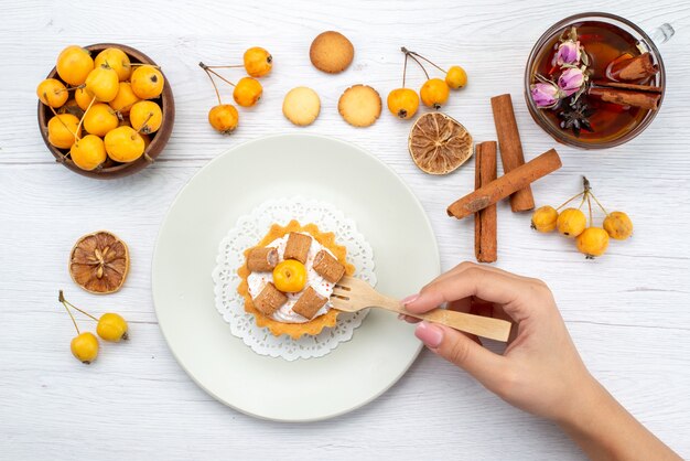 Vista dall'alto della deliziosa piccola torta che viene mangiata da una donna insieme a ciliegie gialle cannella coookies e tè sulla scrivania leggera, biscotto torta biscotto dolce