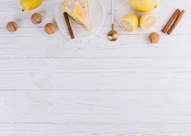 Vista dall&#39;alto della fetta di torta decorata; Limone; Noci; cannella; cucchiaio su sfondo bianco in legno