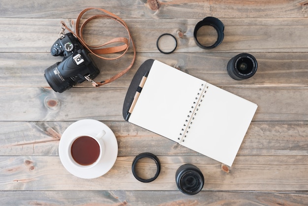 Vista dall&#39;alto della fotocamera dslr; tazza di tè; blocco note a spirale; penna; obiettivo della fotocamera e anelli di prolunga su fondo in legno