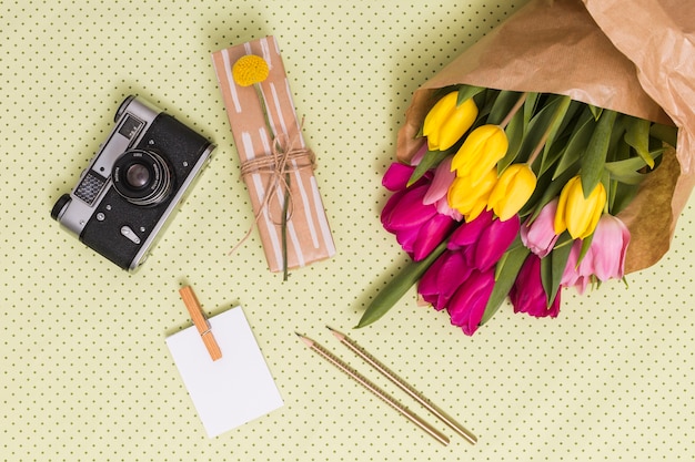 Vista dall&#39;alto della fotocamera retrò; foglio bianco; matite; confezione regalo e bouquet di fiori di tulipano sopra sfondo giallo a pois