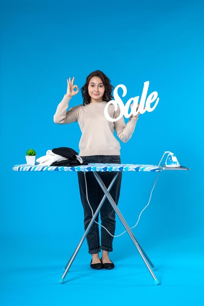 Vista dall'alto della giovane donna in piedi dietro l'asse da stiro e che mostra l'icona di vendita facendo il gesto degli occhiali su sfondo blu