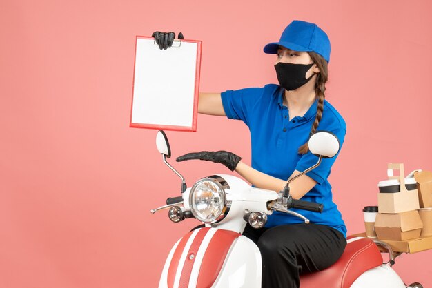 Vista dall'alto della laboriosa ragazza del corriere che indossa maschera medica e guanti seduta su uno scooter con in mano un foglio di carta vuoto che consegna ordini su sfondo color pesca pastello