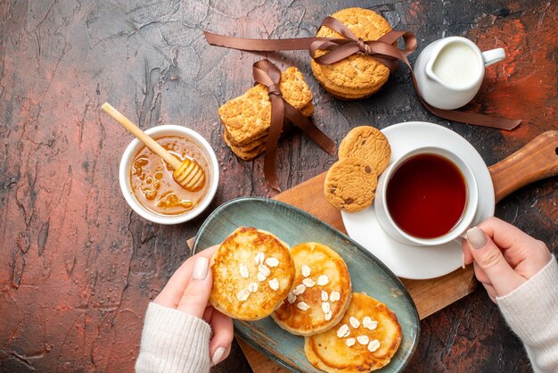 Vista dall'alto della mano che prende il vassoio con frittelle fresche una tazza di tè nero su un tagliere di legno biscotti impilati miele latte su una superficie scura