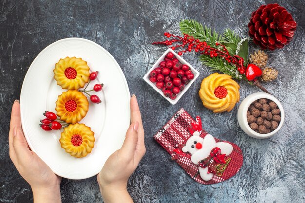 Vista dall'alto della mano che tiene un piatto bianco con deliziosi biscotti decorazione accessorio calza di babbo natale e cornell in una ciotola rami di abete su superficie scura