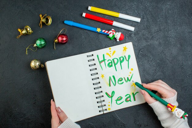 Vista dall'alto della mano che tiene una penna sul taccuino a spirale con accessori di decorazione di scrittura di felice anno nuovo su sfondo nero