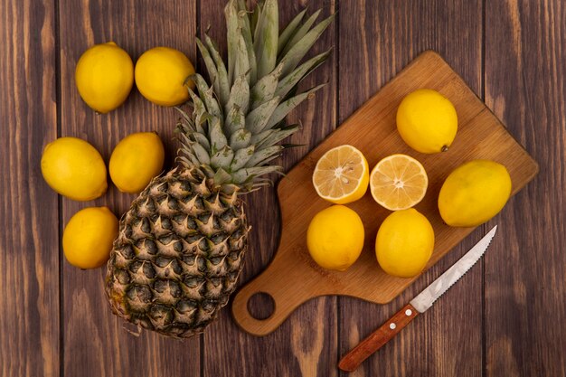 Vista dall'alto della metà fresca e dei limoni interi su una tavola da cucina in legno con coltello con ananas isolato su una parete in legno