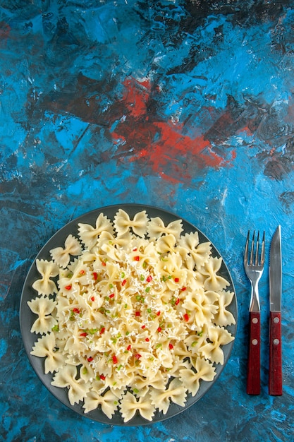 Vista dall'alto della pasta italiana cruda di farfalle con verdure e posate su sfondo blu