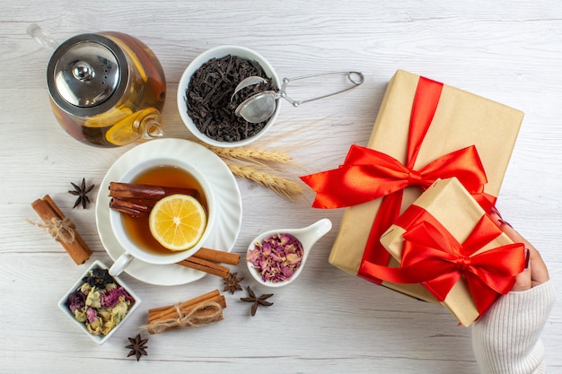 Vista dall'alto della pausa tè con cannella lime e limone in una tazza e una pentola intorno agli ingredienti necessari accanto ai regali con nastro rosso su sfondo bianco