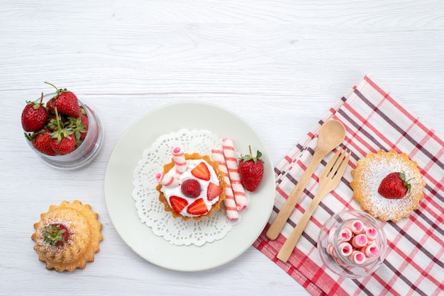 Vista dall'alto della piccola torta con crema e fragole a fette torte caramelle sulla scrivania bianca, zucchero dolce bacca torta di frutta