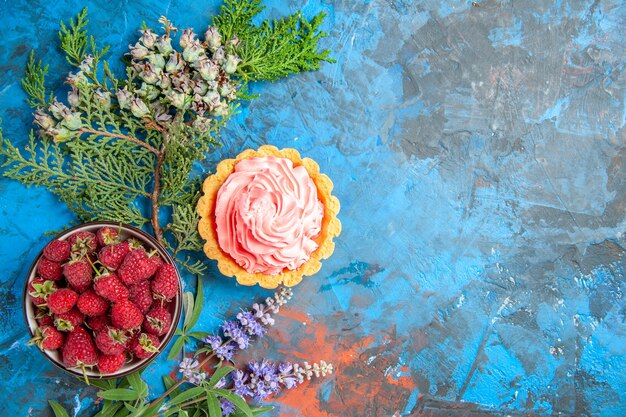 Vista dall'alto della piccola torta con crema pasticcera rosa ciotola con lamponi sulla superficie blu