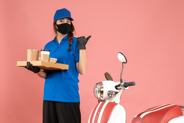 Vista dall'alto della ragazza del corriere che indossa guanti con maschera medica in piedi accanto alla moto con in mano piccole torte di caffè che puntano indietro su sfondo color pesca pastello pastel