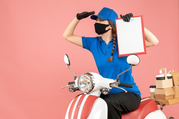 Vista dall'alto della ragazza del corriere concentrato che indossa maschera medica e guanti seduto su uno scooter con un foglio di carta vuoto che consegna ordini su sfondo color pesca pastello