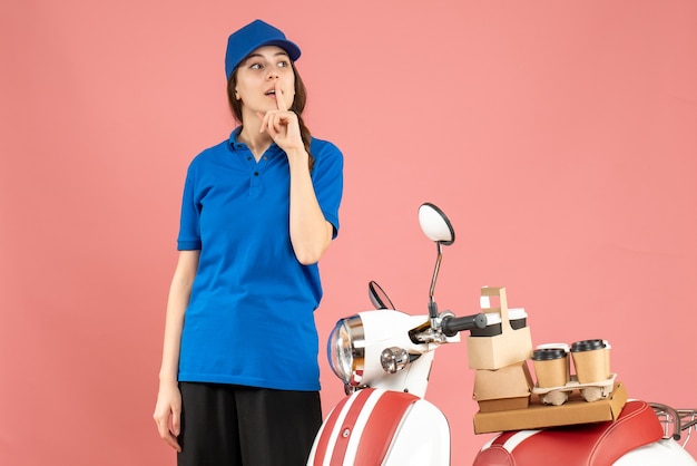 Vista dall'alto della signora del corriere in piedi accanto alla motocicletta con caffè e piccole torte e che fa un gesto di silenzio su uno sfondo color pesca pastello