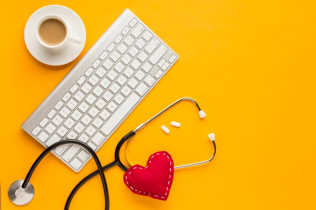 Vista dall'alto della tastiera wireless; compresse; tazza di caffè; stetoscopio; cuore giocattolo cucito; sopra lo sfondo giallo