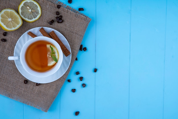 Vista dall'alto della tazza di tè con fetta di limone e cannella sul piattino con fette di limone e pezzi di cioccolato vestirono su sfondo blu con spazio di copia