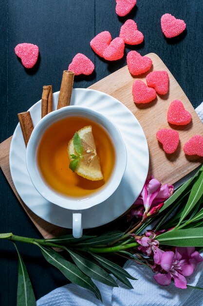 Vista dall'alto della tazza di tè con fetta di limone e cannella sul piattino e marmellate sul tagliere con fiori e foglie con panno su sfondo blu