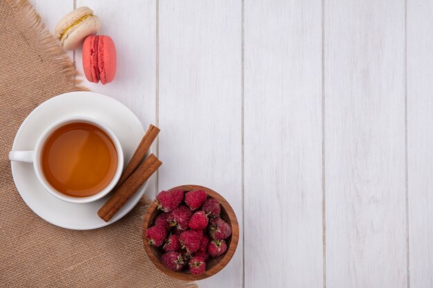 Vista dall'alto della tazza di tè con macarons alla cannella e lamponi su una superficie bianca