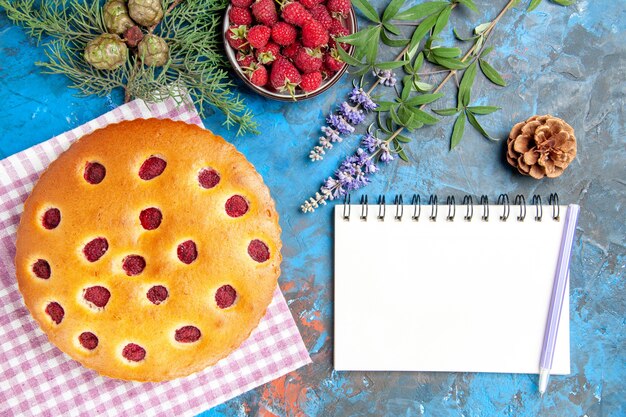 Vista dall'alto della torta di lamponi sulla ciotola del tovagliolo di cucina con il ramo di un albero di pino di lamponi una penna un taccuino sulla superficie blu