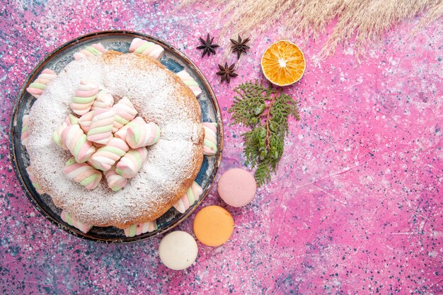 Vista dall'alto della torta di zucchero a velo con marshmallow dolci e macarons sulla superficie rosa