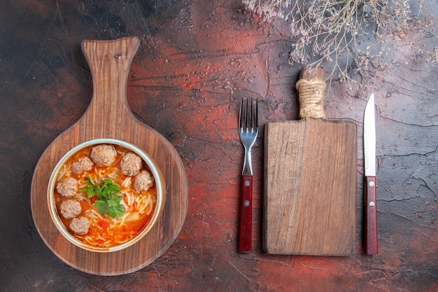 Vista dall'alto della zuppa di polpette di pomodoro con noodles in una ciotola marrone e tagliere con forchetta e coltello su sfondo scuro