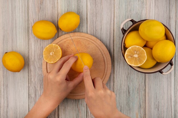 Vista dall'alto delle mani femminili che tagliano il limone su una tavola da cucina in legno con il coltello con i limoni su una ciotola con i limoni isolata su una superficie di legno grigia