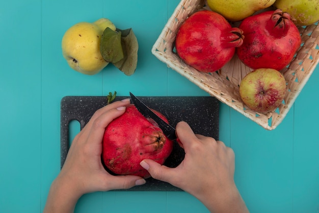 Vista dall'alto delle mani femminili che tagliano il melograno fresco su una tavola di cucina nera con coltello su sfondo blu