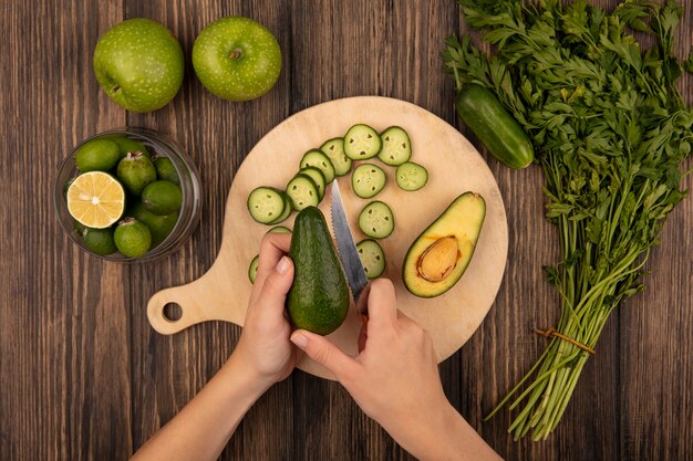 Vista dall'alto delle mani femminili che tagliano un avocado fresco con il coltello su una tavola da cucina in legno con feijoas su una ciotola di vetro con mele verdi e prezzemolo isolato su uno sfondo di legno