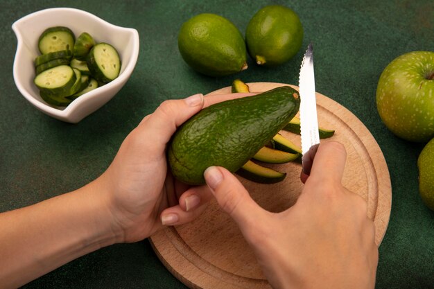 Vista dall'alto delle mani femminili che tagliano un avocado verde brillante con il coltello su una tavola da cucina in legno con limette e mele verdi isolate su una parete verde