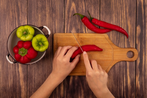 Vista dall'alto delle mani femminili che tagliano un peperoncino su una tavola di cucina in legno con coltello su una parete in legno