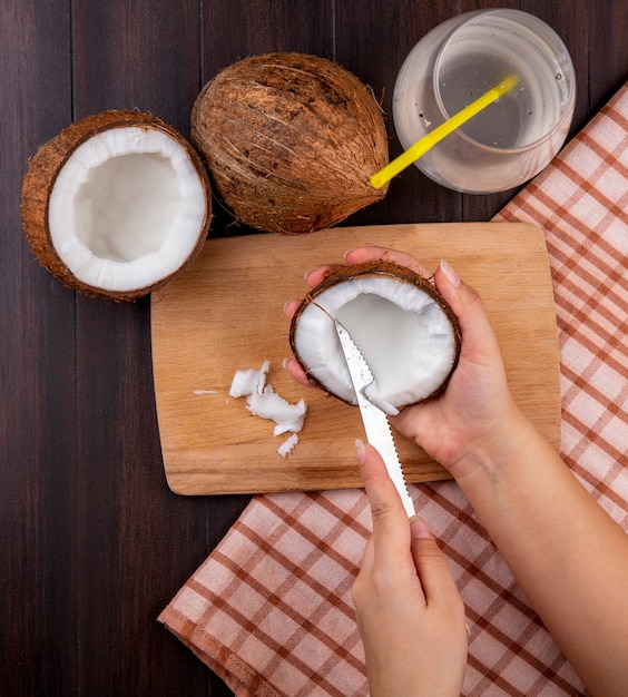 Vista dall'alto delle mani femminili che tengono il cocco in una mano e il taglio nell'altra mano sul bordo della cucina in legno con noci di cocco e un bicchiere d'acqua sulla tovaglia a quadri sul nero