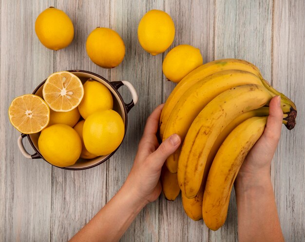 Vista dall'alto delle mani femminili che tengono le banane con i limoni su una ciotola con i limoni isolati su un fondo di legno grigio