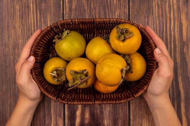 Vista dall'alto delle mani femminili che tengono un secchio di cachi arancioni freschi frutti su un tavolo di legno