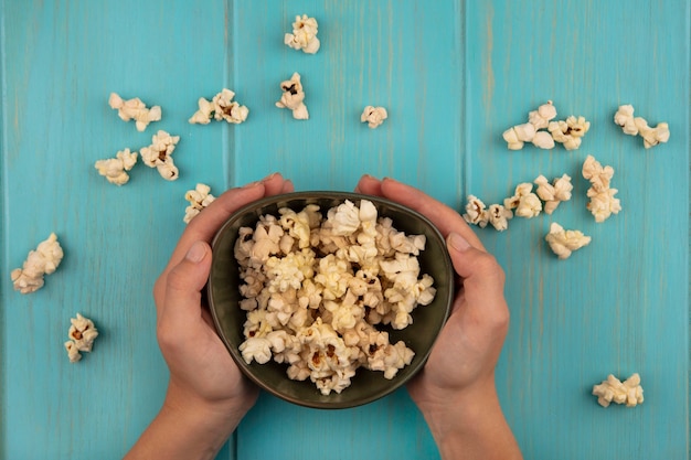 Vista dall'alto delle mani femminili che tengono una ciotola di gustosi popcorn con popcorn isolati su un tavolo di legno blu