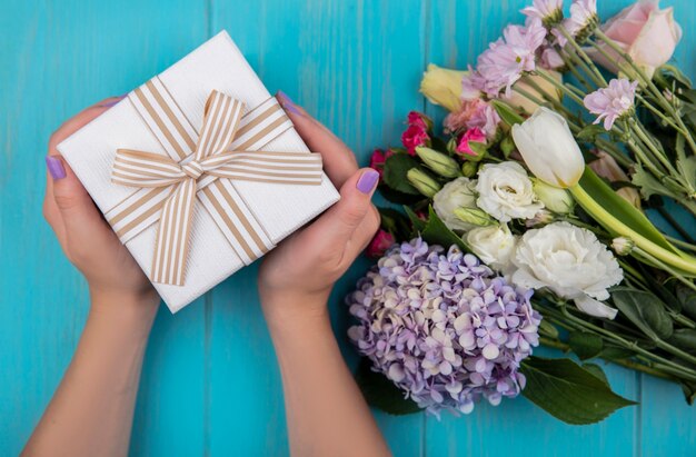 Vista dall'alto delle mani femminili che tengono una confezione regalo con meravigliosi fiori freschi come la rosa gardenzia tulip daisy isolata su uno sfondo di legno blu