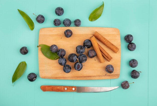 Vista dall'alto delle piccole prugnole di frutta nerastre acide su una tavola di cucina in legno con bastoncini di cannella con coltello su sfondo blu