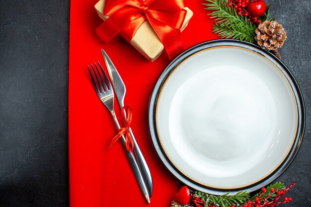 Vista dall'alto dello sfondo di Capodanno con posate piatto da pranzo set decorazione accessori rami di abete accanto a un regalo su un tovagliolo rosso