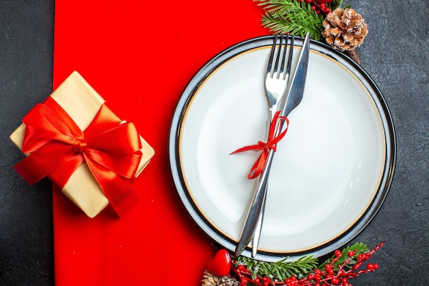 Vista dall'alto dello sfondo di Natale con posate con nastro rosso su un piatto da pranzo accessori per la decorazione rami di abete accanto a un regalo su un tovagliolo rosso