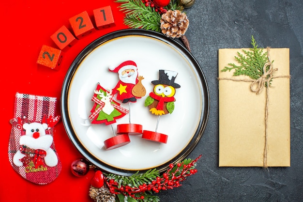 Vista dall'alto di accessori per la decorazione del piatto da cena rami di abete e numeri calza di Natale su un tovagliolo rosso accanto al regalo su una tavola nera