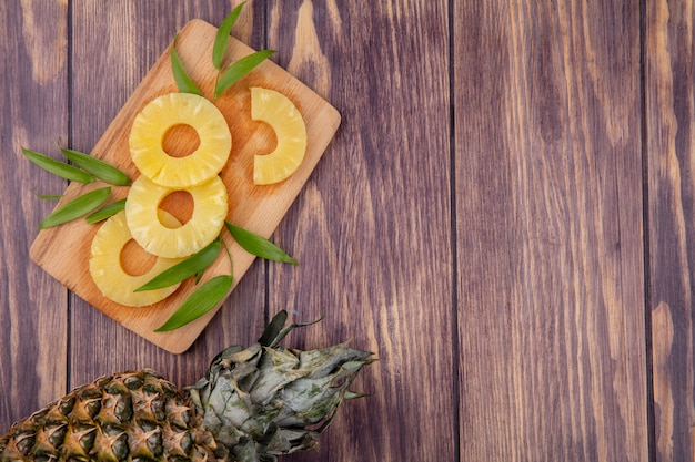 Vista dall'alto di ananas e fette con foglie sul tagliere e superficie in legno