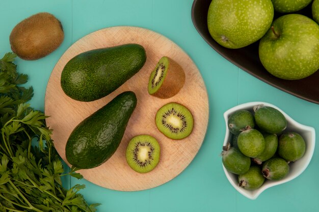 Vista dall'alto di avocado con fette di kiwi su una tavola da cucina in legno con feijoas su una ciotola con le mele su una ciotola su una parete blu