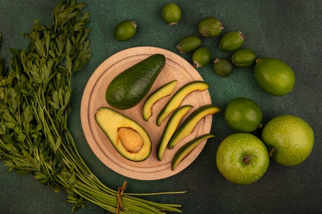 Vista dall'alto di avocado dalla pelle verde con fette su una tavola da cucina in legno con coltello con lime, mele verdi, feijoas e prezzemolo isolato su una superficie verde