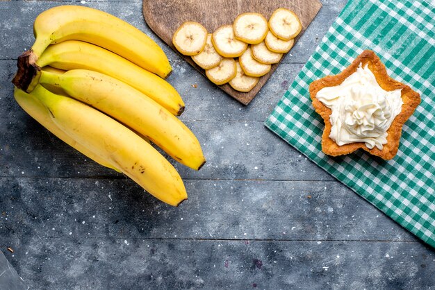 vista dall'alto di bacche intere di banane gialle fresche con torta sulla scrivania grigia, gusto di vitamina bacche di frutta