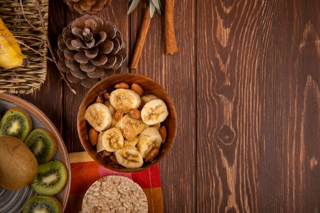 Vista dall'alto di banane a fette con mandorle in una ciotola di legno, fette di kiwi su un piatto e cracker di riso su rustico con spazio di copia
