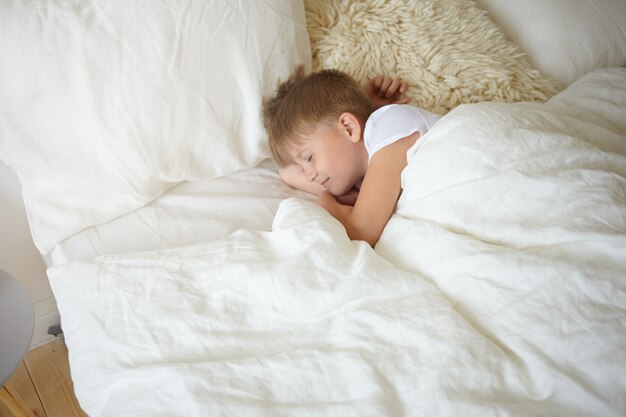 Vista dall'alto di bello scolaro adorabile di aspetto europeo che ha un pisolino dopo le lezioni a scuola. Dolce ragazzo affascinante in maglietta bianca che dorme pacificamente nel letto su lenzuola bianche, sorridente addormentato