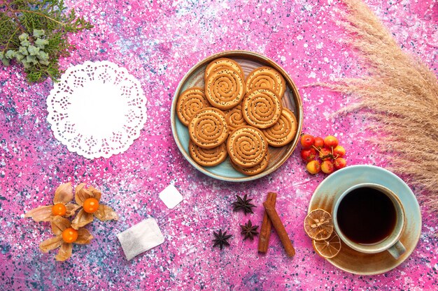 Vista dall'alto di biscotti dolci all'interno del piatto con cannella e tazza di tè sulla superficie rosa