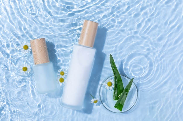 Vista dall'alto di bottiglie per la cura della pelle su una superficie d'acqua azzurra con aloe vera e fiori di margherita